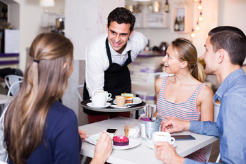 young smiling waiter bringing ordered dishes to friends in tearoom