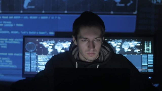 Geek Hacker programmer is working on computer in cyber security center filled with display screens.