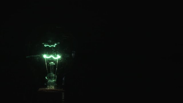 Green light bulb framed left with empty black space framed right, turns on and off with flicker, close up.