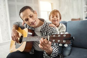 Relaxing together. Attractive alert dark-haired man smiling and playing the guitar and his son hugging him