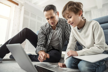 Searching the net. Good-looking inspired dark-haired man smiling and looking at the laptop and his son sitting near him on the floor