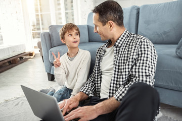 Best friends. Handsome alert fair-haired boy smiling and sitting on the floor with his father and his daddy holding a laptop and they looking at each other