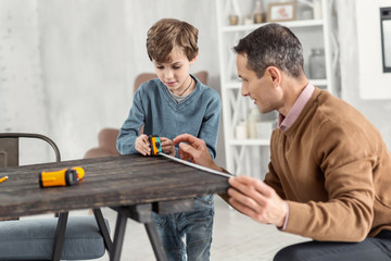 Measuring. Good-looking concentrated little fair-haired boy holding a measuring tape and measuring the table and his daddy helping help