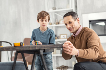 Happy family. Nice content little fair-haired boy holding a measuring tape and measuring the table and his daddy helping help