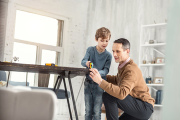 Caring daddy. Handsome serious little fair-haired boy holding a measuring tape and measuring the table and his daddy helping help