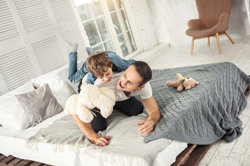 Our day off. Attractive inspired dark-haired daddy laughing and lying on the bed with his son and they having fun