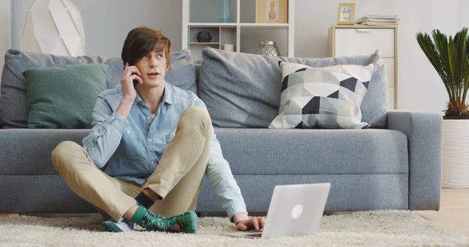 Young Caucasian man in the casual style working at the laptop computer and calling on the telephone while sitting on the floor in the nice comfortable living room. Indoor