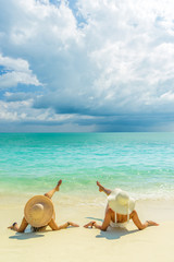 Fototapeta na wymiar Sexy bikini body of two women enjoy the sea by laying down on sand of beach wearing hat. Happy island lifestyle. White sand and crystal sea of tropical beach.