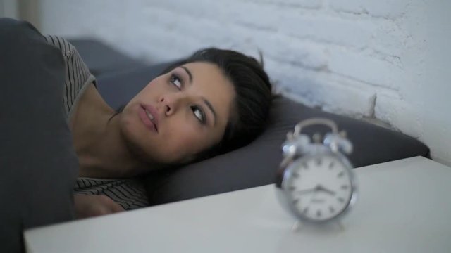 lateral panning shot of young attractive hispanic woman upset in stress and insomnia lying anxious on bed trying to sleep suffering sleeping disorder at night looking alarm clock frustrated 