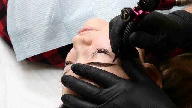 Beauty salon. Close up of Male beautician in black gloves making permanent makeup procedure on female eyebrows. Young woman gets facial beauty procedure. Facial rejuvenation. Using tattoo machine