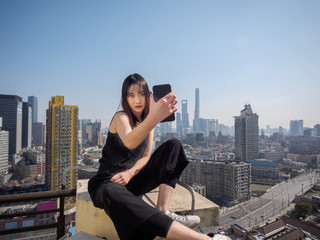 Fototapeta premium Beautiful young brunette woman in black dress sit on top of mansion roof and take selfie photo with blur Shanghai Bund landmark buildings background. Emotions, people, beauty and lifestyle concept.