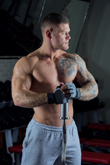 Muscular shirtless man with blue eyes and tattoo poses with barbell in gloves and gray pants