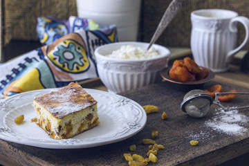 Cottage cheese casserole with raisins and dried apricots. A piece of casserole with powdered sugar on a white plate, fresh farmer cottage cheese in a white bowl on a wooden table. Rustic style.