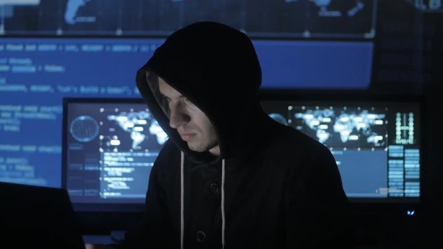 Man geek hacker in hood working at computer while blue code characters reflect on his face in cyber security center filled with display screens.