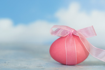pink painted easter egg with a ribbon against a blurry blue sky with white clouds, copy space