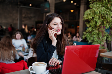 Brunette girl sitting on cafe, working with red laptop and speaking at mobile phone.