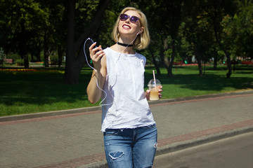 young sunny hot girl in white blouse , jeans and glasses standing smiling and listening music from mobile phone in one hand with ice coffee in other hand