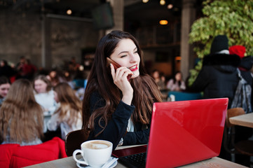 Brunette girl sitting on cafe, working with red laptop and speaking at mobile phone.