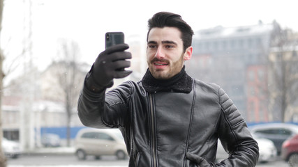 Handsome trendy man wearing black leather jacket using cell phone to take photos, outdoor in city setting in winter day shot