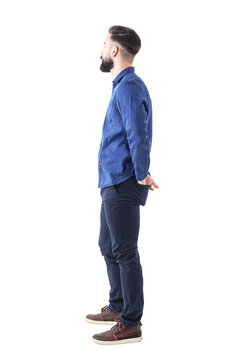 Side view of young stylish bearded man with hands in back pockets standing and watching. Full body isolated on white background. 