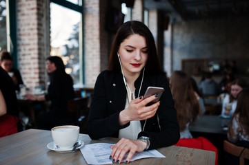 Brunette girl sitting on cafe with cup of cappuccino, listening music on headphones and looking at mobile phone.