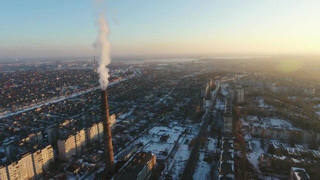 A bird`s eye view of a high cooling tube with dense swhite moke between multistoried buildings at sunset in winter. The drone flies away 
