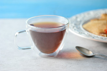 Glass double-walled cup. In the cup black tea. In the frame, a teaspoon and the edge of the plate. Light background. Close-up.