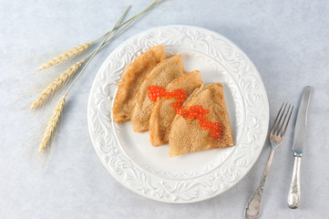 Pancakes thin on a white plate. On the pancakes is red caviar. Next to the plate are wheat ears and cutlery. Light background. View from above. Close-up. Macro photography.