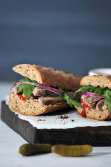 Sandwich on a wooden board. The filling of the sandwich consists of beef, arugula, red onion and...