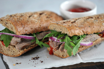 Sandwich on a wooden board. The filling of the sandwich consists of beef, arugula, red onion and marinated cucumber slices. In the frame,  a cup of ketchup.  Close-up. Macro photography.