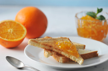 Orange jam and plate with toast. In the frame, an orange and half an orange. One toast is smeared with jam. Next to the plate is a teaspoon. Light background. Close-up. Macro photography.