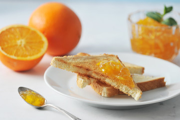 Orange jam and plate with toast. In the frame, an orange and half an orange. One toast is smeared with jam. Next to the plate is a teaspoon with jam. Light background. Close-up. Macro photography.