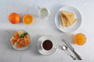 Table setting for breakfast. In the frame orange jam, toast, tea, milk in the milkman, marmalade in the form of orange slices, oranges, cutlery. Light background. Close-up. View from above.