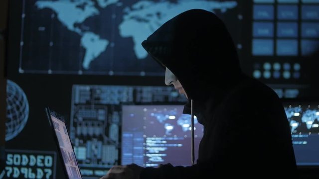 Man geek hacker in hood working at computer in cyber security center filled with display screens.
