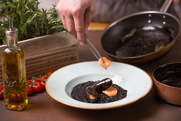 The side view of the chef cooking the seafood salad consisted of the black rice, shrimps, mussels placed on the white plate.