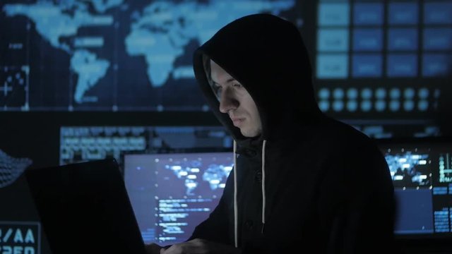 Man geek hacker in hood working at computer in cyber security center filled with display screens.