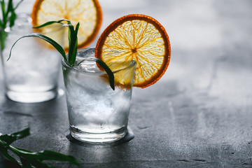 Two shots of cocktails with ice, dried orange slice and fresh tarragon on a grey table.
