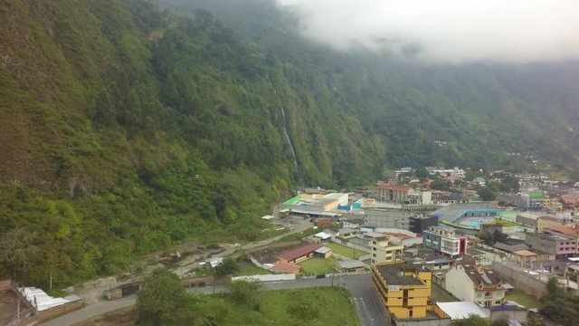 An aerial shot of the town of Banos de Agua Santa, Ecuador, built along a river at the base of a steep verdant mountain, a thin waterfall down the cliff and low clouds over the peaks.