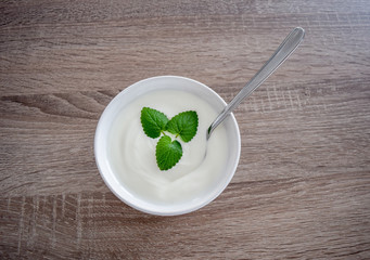 Plain yogurt in a ceramic bowl with mint leaves as decoration and spoon inside the yogurt, isolated on a wooden table from top vie, white yoghurt