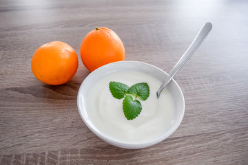 Plain yogurt with mint leaves and spoon inside a ceramic bowl, oranges as decoration on a wooden background from above, white yoghurt