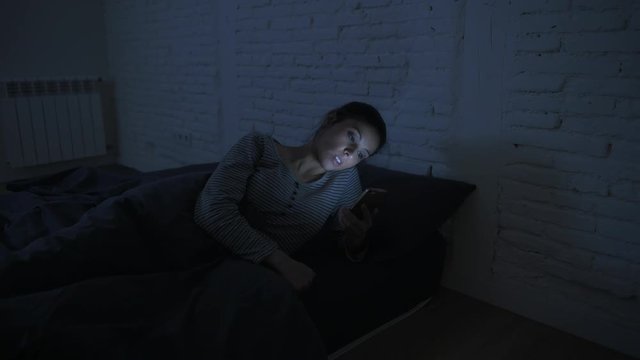 low light video lateral panning shot of young attractive hispanic woman on her 30s lying in bed late night using mobile phone looking tired and bored in internet addiction concept 
