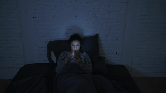 low light video frontal shot of young attractive hispanic woman on her 30s lying in bed late night using mobile phone looking tired and bored in internet addiction concept