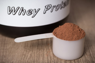Whey Protein. White scoop with chocolate flavour powder and black jar. Wooden table. Close up, texture