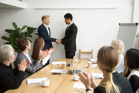 African boss handshaking caucasian subordinate congratulating with promotion, appreciating good work result, team supporting applauding black ceo and white employee shaking hands at group meeting