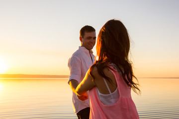 Fototapeta premium Young couple is holding hands and spinning in the water on summer beach. Sunset over the sea. Just married couple hugging. Romantic love story. Man and woman in holiday honeymoon trip. Happy emotions.