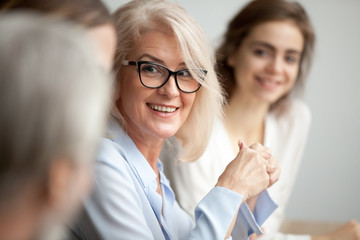 Smiling aged businesswoman in glasses looking at colleague at team meeting, happy attentive female team leader listening to new project idea, coach mentor teacher excited by interesting discussion - obrazy, fototapety, plakaty