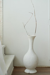 White vase with dry branch stands in front of a white wall. The minimalistic design of the room