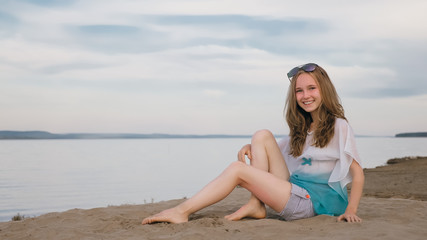 One beautiful teenage girl with brown hair outside on a beautiful summer day. The woman is sitting on the beach. The sun was gone in the sunset. The girl is genuinely genuinely genuinely smiling and