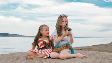 Two teenage are sitting on a sandy beach. They wear sunglasses. Sisters talk to each other and selfie on phone. They have two dogs. Girls correspond, playing, on the Internet.