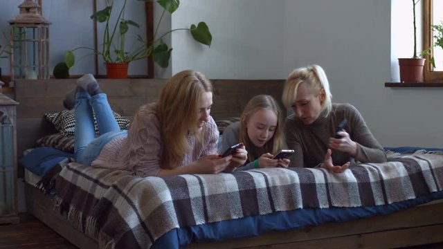 Joyful attractive women and little girl surfing the net on mobile phones, sharing media content on line on smart phones and chatting while lying on the bed in bedroom during spending leisure at home.